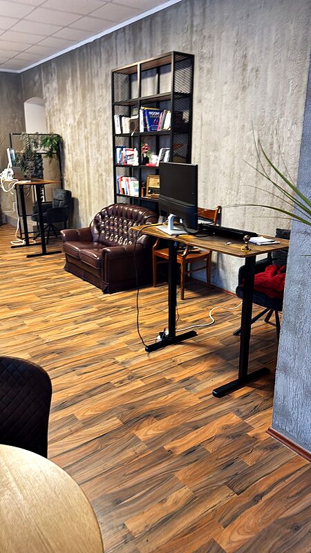Höhenverstellbarer Schreibtisch mit Monitor vor einem Bücherregal und dem Ledersofa, dahinter ein zweiter Arbeitsplatz.