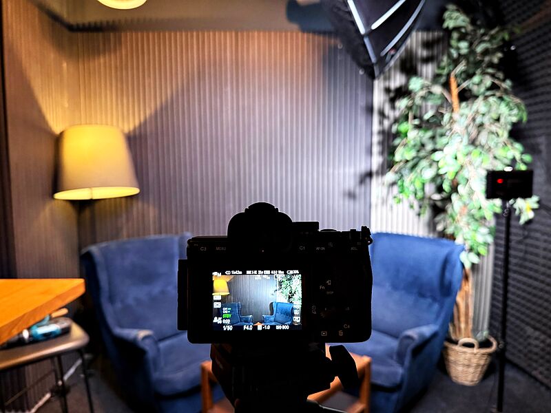 Blick über die Sony-Kamera auf zwei blaue Ohrensessel vor den Akustiklamellen, daneben Stehlampe, Pflanze und Studioleuchte.
